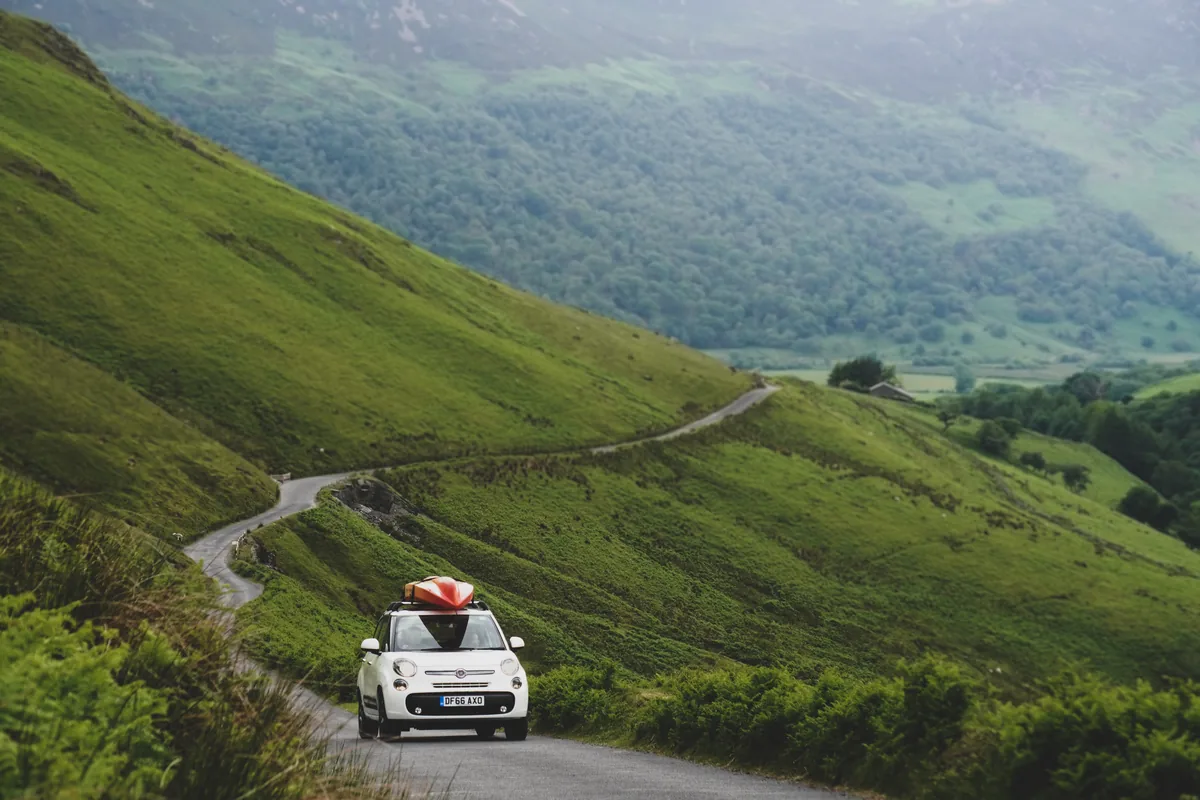 A white car with a red kayak on its roof on a road winding through green hills