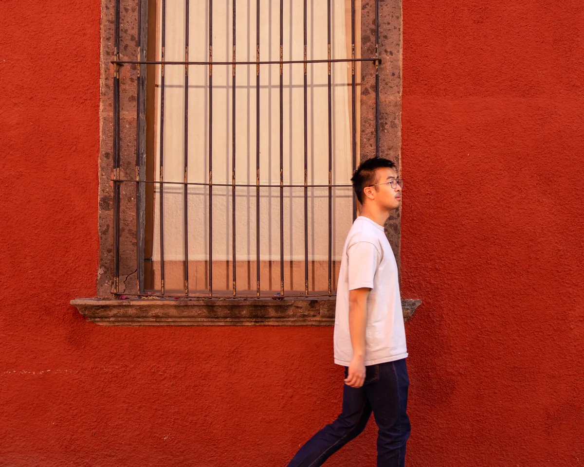 A man in a white T-shirt walking past a barred window in a red wall
